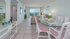 Habitación de hotel moderna con una mesa de comedor de vidrio, sillas de mimbre blanco, un sofá y vista al océano.