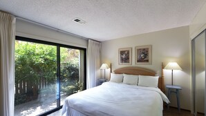 A hotel room with a large bed, two bedside lamps, a view of greenery through a sliding door, and two framed pictures on the wall.