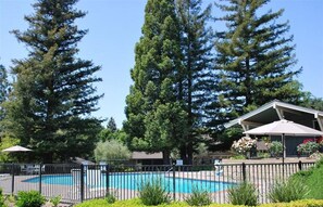 A swimming pool surrounded by a fence, with tall trees in the background and a building with an umbrella.
