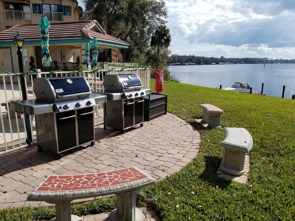 An outdoor patio with two barbecue grills, a stone table, and a view of a lake.