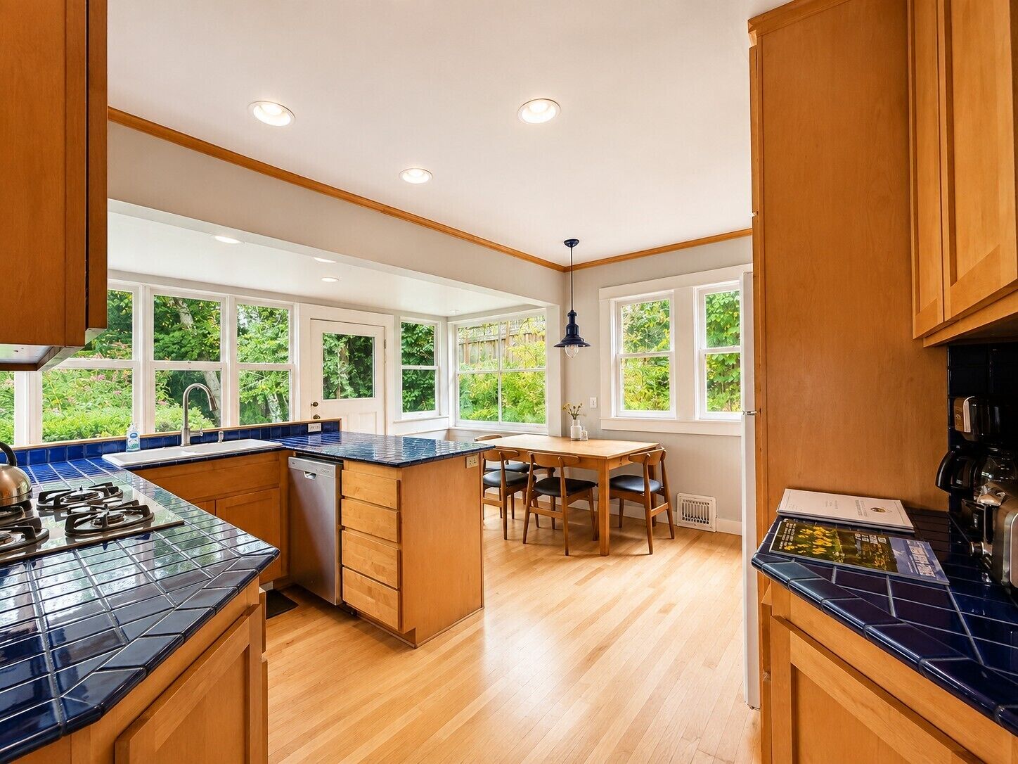 A kitchen with wooden cabinets, a blue tiled countertop, a dining table with chairs, and large windows with a view of greenery.