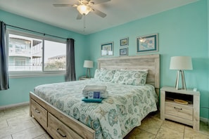 Quarto com cama, mesa de cabeceira, abajur, ventoinha no teto e janela com cortinas.