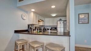 Una cocina con armarios blancos, un refrigerador de acero inoxidable, un microondas y una cafetera. Hay tres taburetes de bar con asientos acolchados.