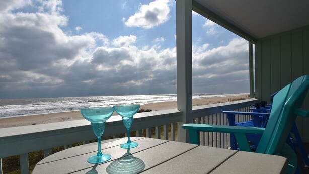 Una mesa con dos vasos azules en una terraza de madera con vistas a una playa y olas.