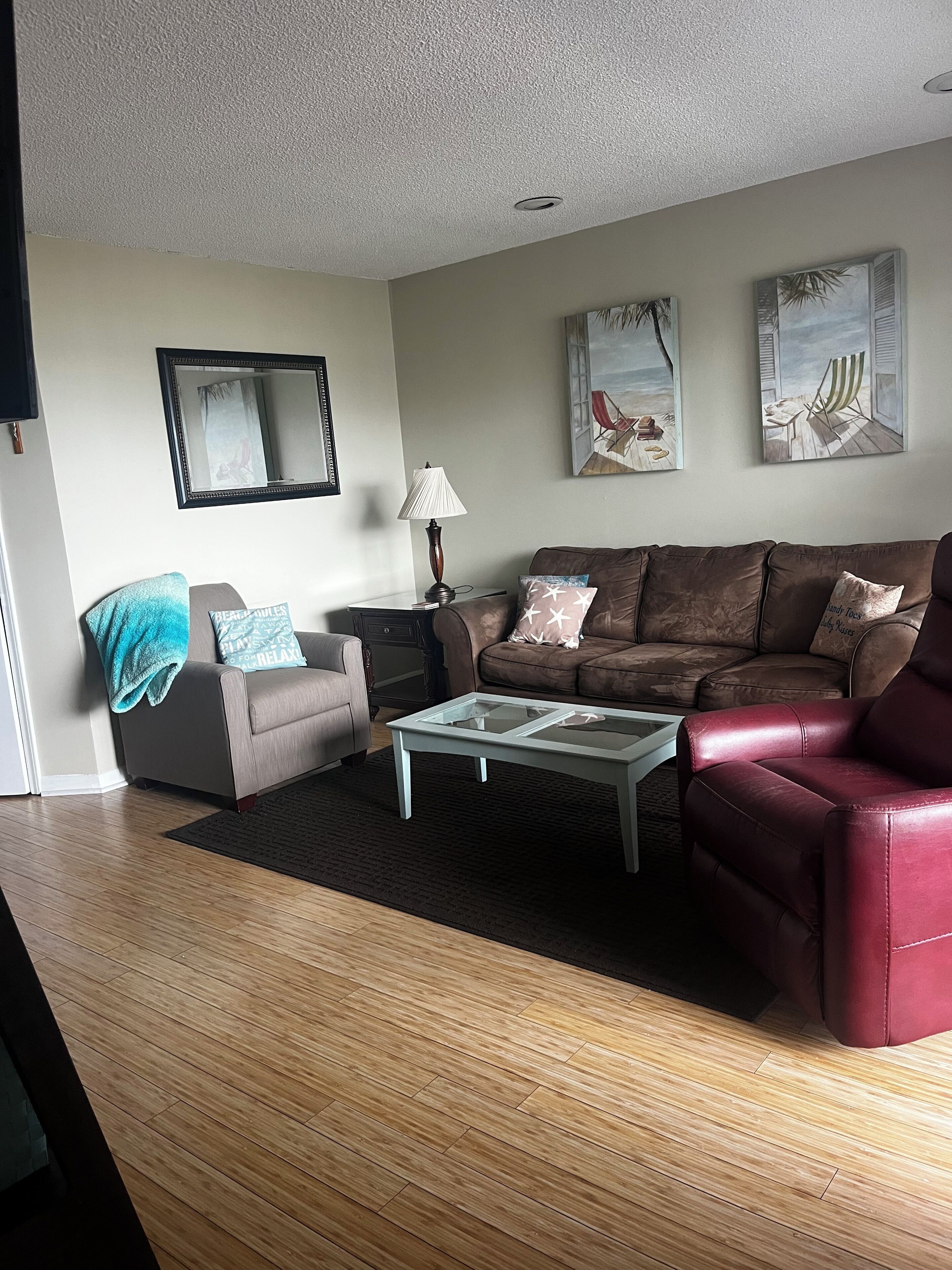 A living room with a brown sofa, a red armchair, a glass coffee table, a lamp, and two wall-mounted paintings.