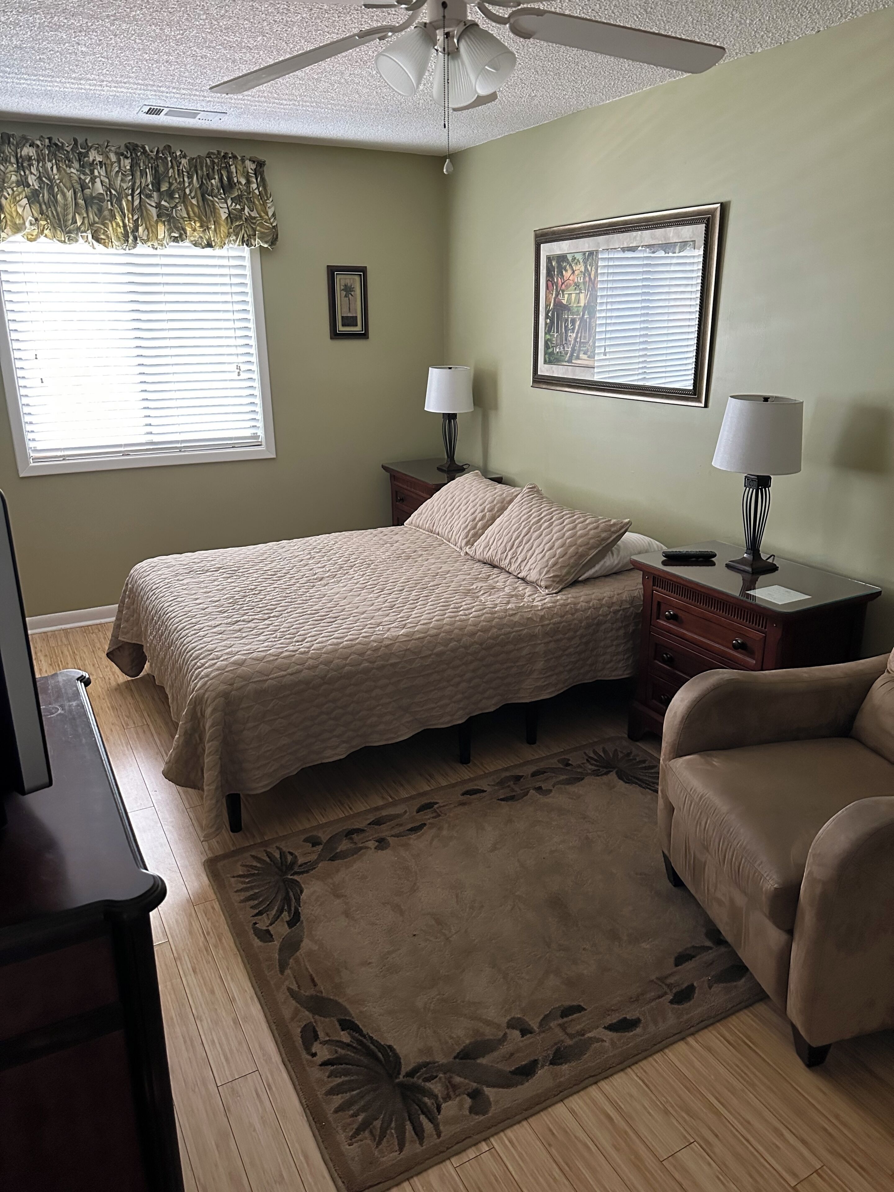 A bedroom with a bed, two bedside tables with lamps, a ceiling fan, a window with blinds, a framed picture on the wall, and a floral rug.