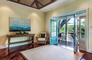 A room with a wooden floor, a white rug, a chair, a table with a vase of flowers, and a large window with a view of greenery. - Luxurious Oceanfront Beach House w/AC & Stunning Coastal Views: Puako Bay (74) (Kamuela)