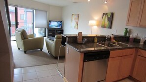 A compact kitchen with a dishwasher, a sink, and a small island. There is a television in the living area with a sofa and chairs. - Waterfront Pearl Home Away from Home (Portland)