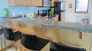 A kitchen with wooden cabinets, a granite countertop, a microwave, a refrigerator, and a window with blinds. - Waterfront Pearl Home Away from Home (Portland)