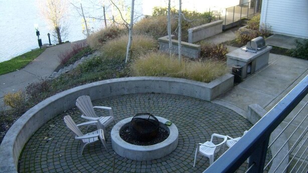A circular outdoor seating area with Adirondack chairs, a fire pit, and a grill, surrounded by a paved walkway and a view of a body of water. - Waterfront Pearl Home Away from Home (Portland)