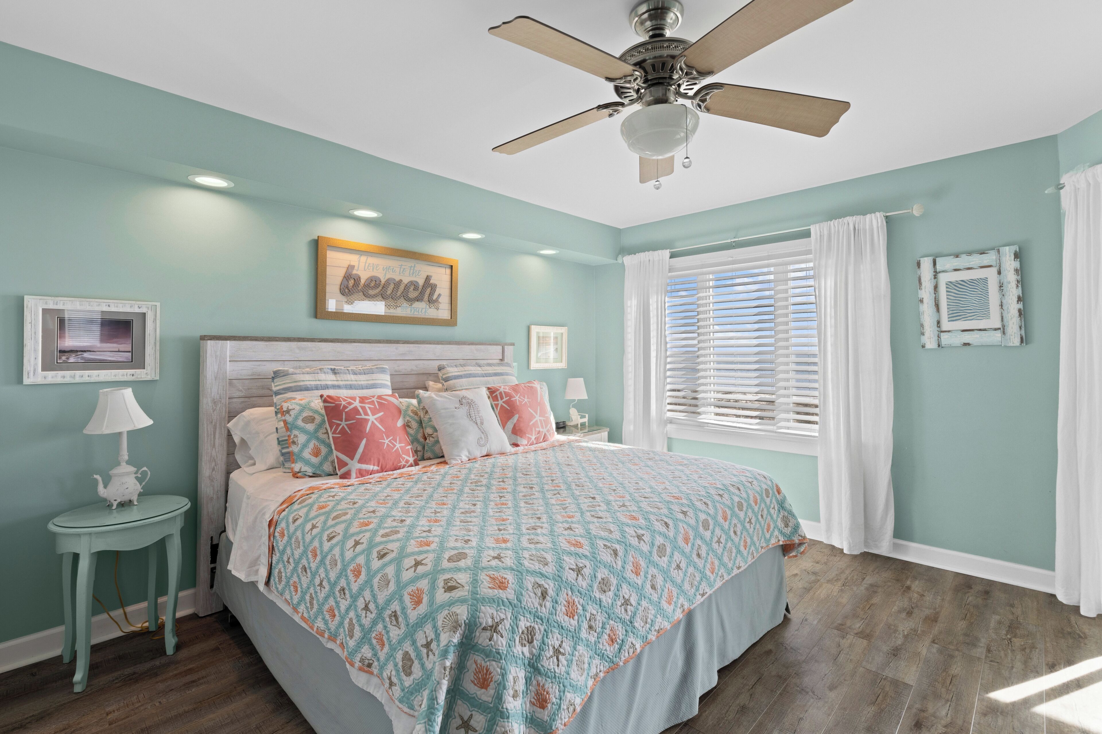 Une chambre à coucher avec un lit, un ventilateur de plafond, une fenêtre avec des stores et un mur décoré de tableaux encadrés.