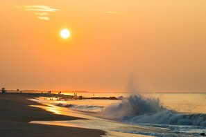 Een strandgezicht met een ondergaande zon boven de oceaan, golven die op het strand breken en een uitkijktoren in de verte.