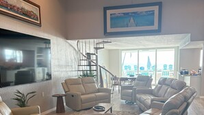 Uma sala de estar espaçosa com uma televisão de tela grande, poltronas de couro, ventilador de teto e vista para o exterior por grandes janelas.