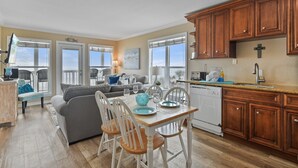 A modern kitchen with wooden cabinets, a dining table with chairs, and a view of the outdoors through large windows. - "Beachfront Paradise - Wake Up to Ocean Views & Waves! (Galveston)