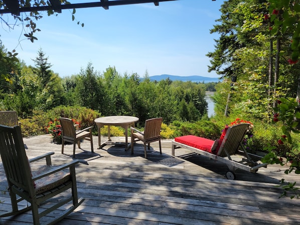 A wooden deck with outdoor furniture, including chairs and a table, overlooking a forested area and a body of water.