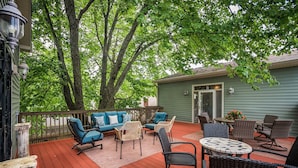 A covered outdoor patio with a red wooden deck, blue and beige wicker furniture, and a green house in the background. - The Highlands Hangout, EXPO, DOWNTOWN, restaurant row!!!  Game room & LOCATION! (Louisville)