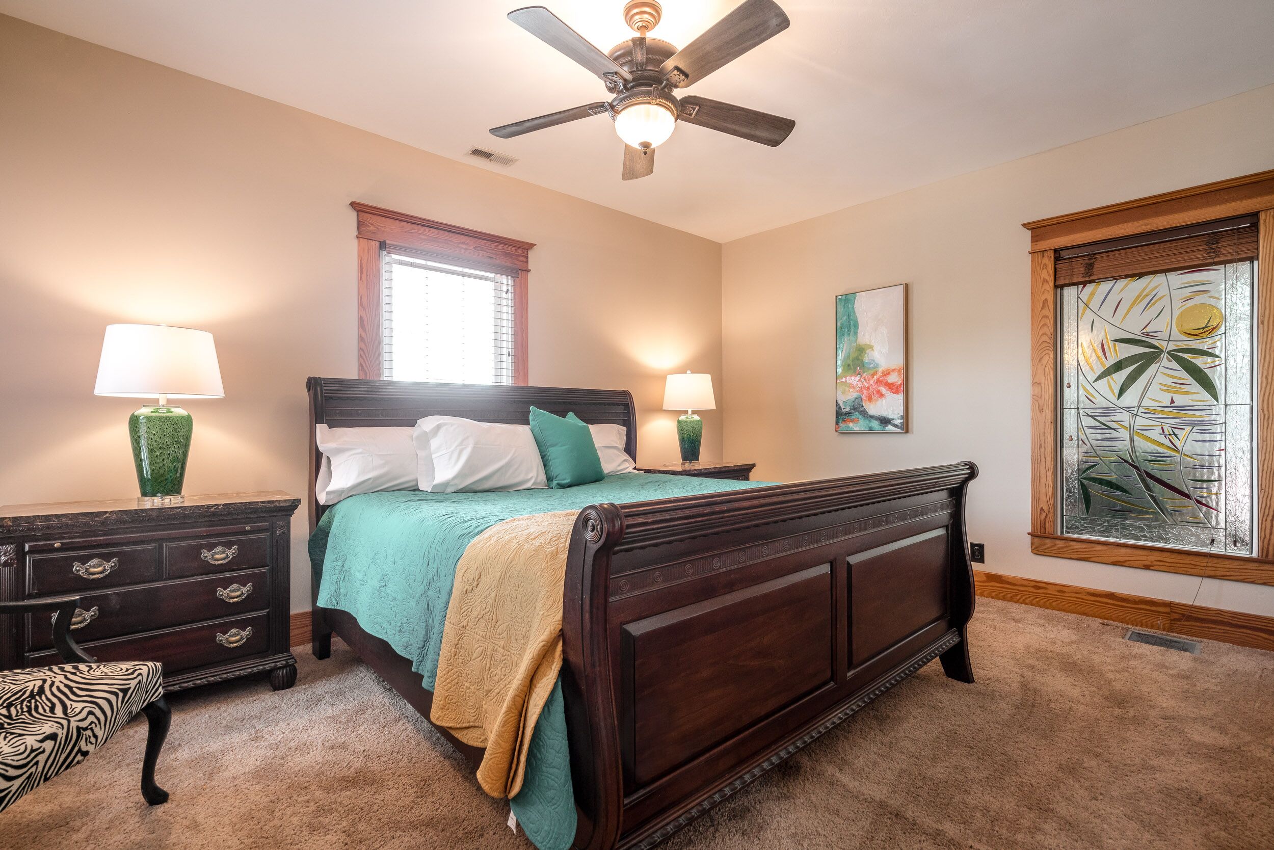 A bedroom with a large bed, two bedside tables, a ceiling fan, a window with blinds, and a decorative glass panel.