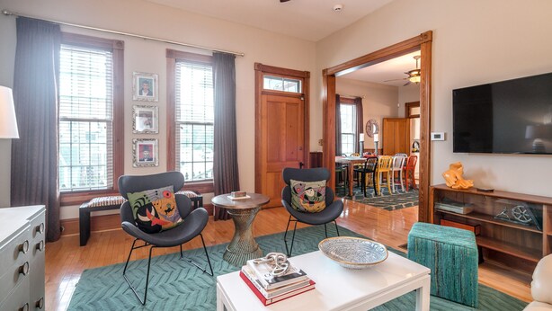 A spacious living room with a large TV, two armchairs with colorful cushions, a coffee table with books, and a dining area in the background. - The Highlands Hangout, EXPO, DOWNTOWN, restaurant row!!!  Game room & LOCATION! (Louisville)