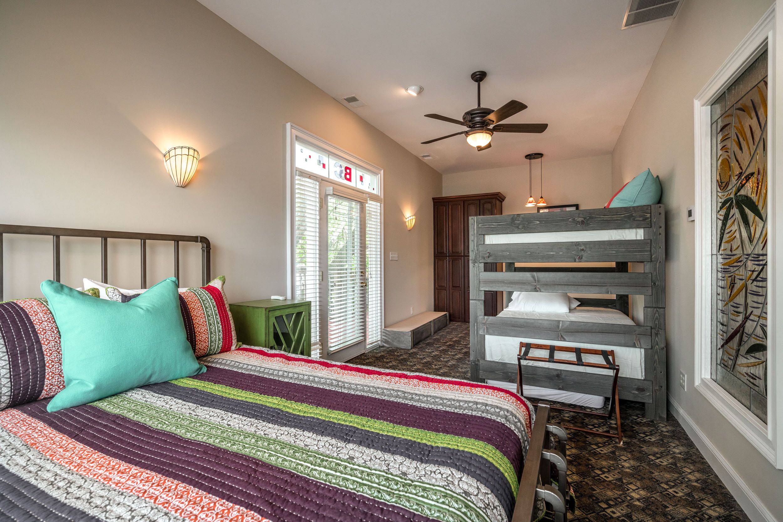 A bedroom with a bed, a bunk bed, a ceiling fan, a wall-mounted light, and a window with blinds.