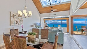 A spacious living area with a high ceiling, a dining table with wicker chairs, a sofa, and a large window offering a view of the ocean.