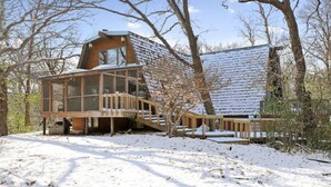 Une cabane en bois dotée d’une véranda en verre, d’une terrasse en bois et d’un toit recouvert de neige.