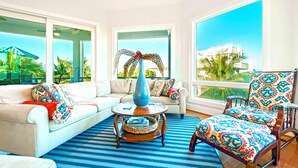 A spacious living room with a white sofa, a patterned armchair, a blue vase on a wooden table, and large windows offering a view of palm trees and a building.