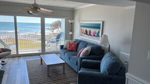 A living room with a view of the ocean, a ceiling fan, a sofa with cushions, a coffee table, and a lamp.