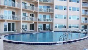 A swimming pool with a 6 ft 6 in depth, surrounded by a tiled deck, and a multi-story building with balconies in the background.