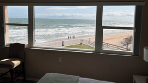 A room with a large window overlooking a beach, a chair, and a bed.