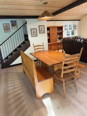 Une table à manger en bois, avec des bancs, un escalier et une bibliothèque, dans une pièce dont le plafond est orné de poutres en bois.