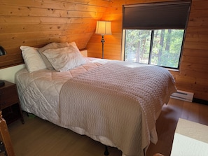Une pièce aux murs lambrissés, comprenant un lit, une table de chevet, une lampe et une fenêtre donnant sur la forêt.