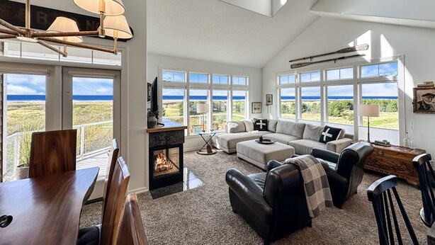 A spacious living room with a fireplace, large windows offering ocean views, and a high ceiling.