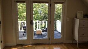 Una stanza con pavimento in legno, un comò bianco e una porta-vetro scorrevole che dà su un patio esterno con ringhiera bianca e una sedia in vimini.