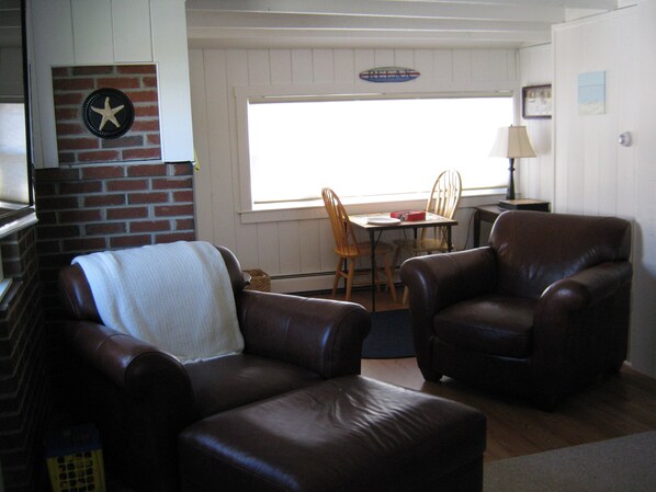 A living room with two leather armchairs, a brick accent wall, a dining table with chairs, and a window with blinds. - Last minute discount. Private beach on Harborside . Sleeps 10. Kayaks included (Wells)