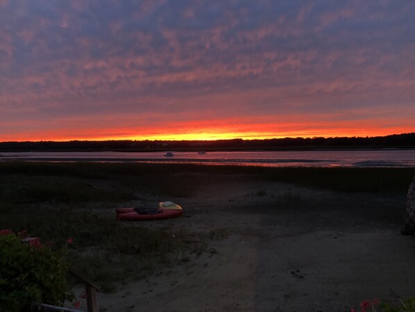 A sunset over a body of water with boats, a red kayak, and a grassy area in the foreground.