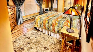 A room with wooden ceiling and walls, a bed with a colorful blanket, a desk with a lamp, and a large window with curtains.