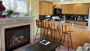 A modern kitchen with wooden cabinets, a built-in island with bar stools, a fireplace, and a large window.