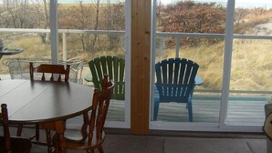 A covered outdoor dining area with a table and chairs, large windows with a view of the beach and trees.