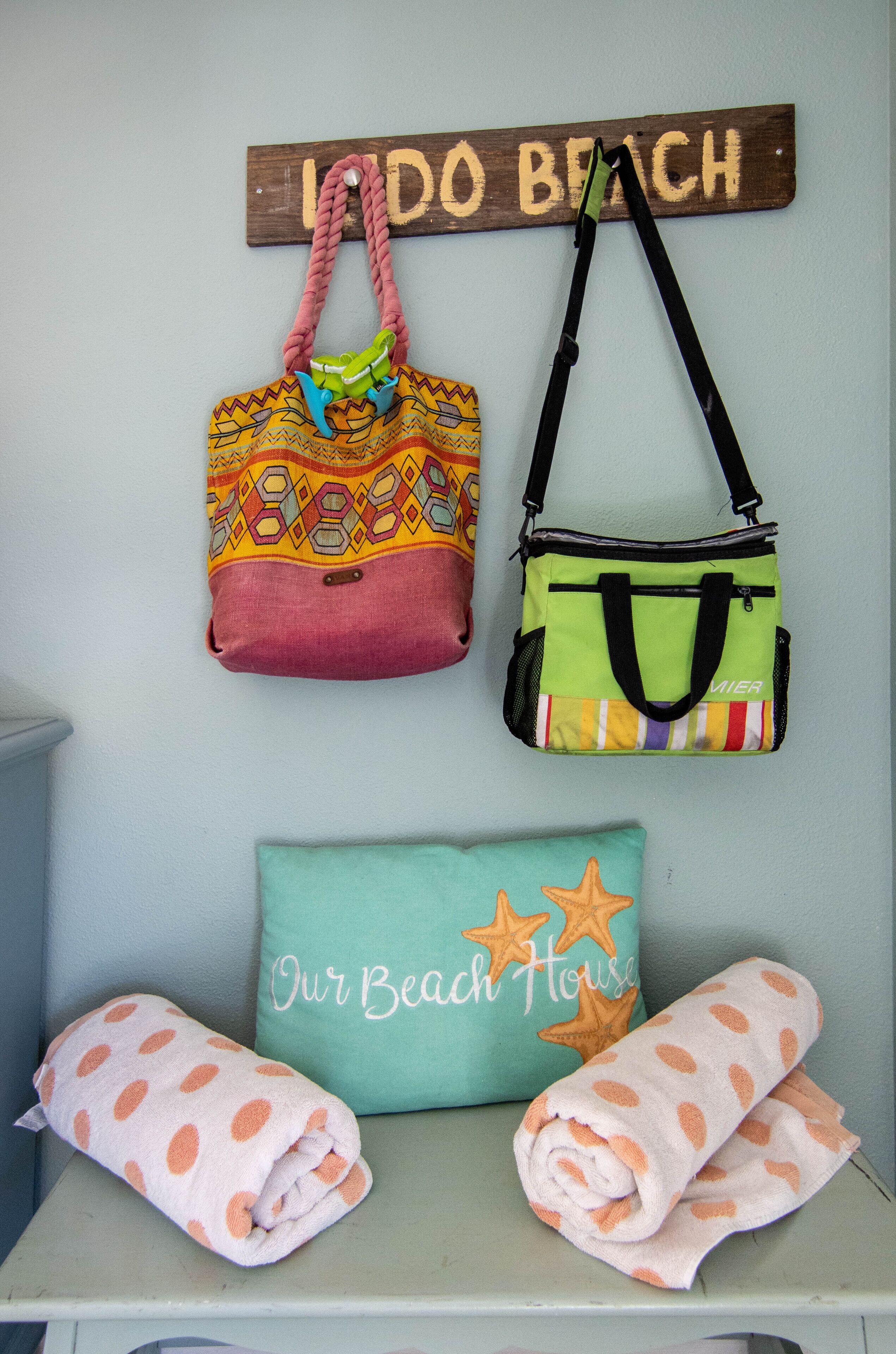 Una pared con un letrero que dice 'LIDO BEACH', dos bolsas coloridas colgadas y una almohada que dice 'Our Beach House' junto con toallas.