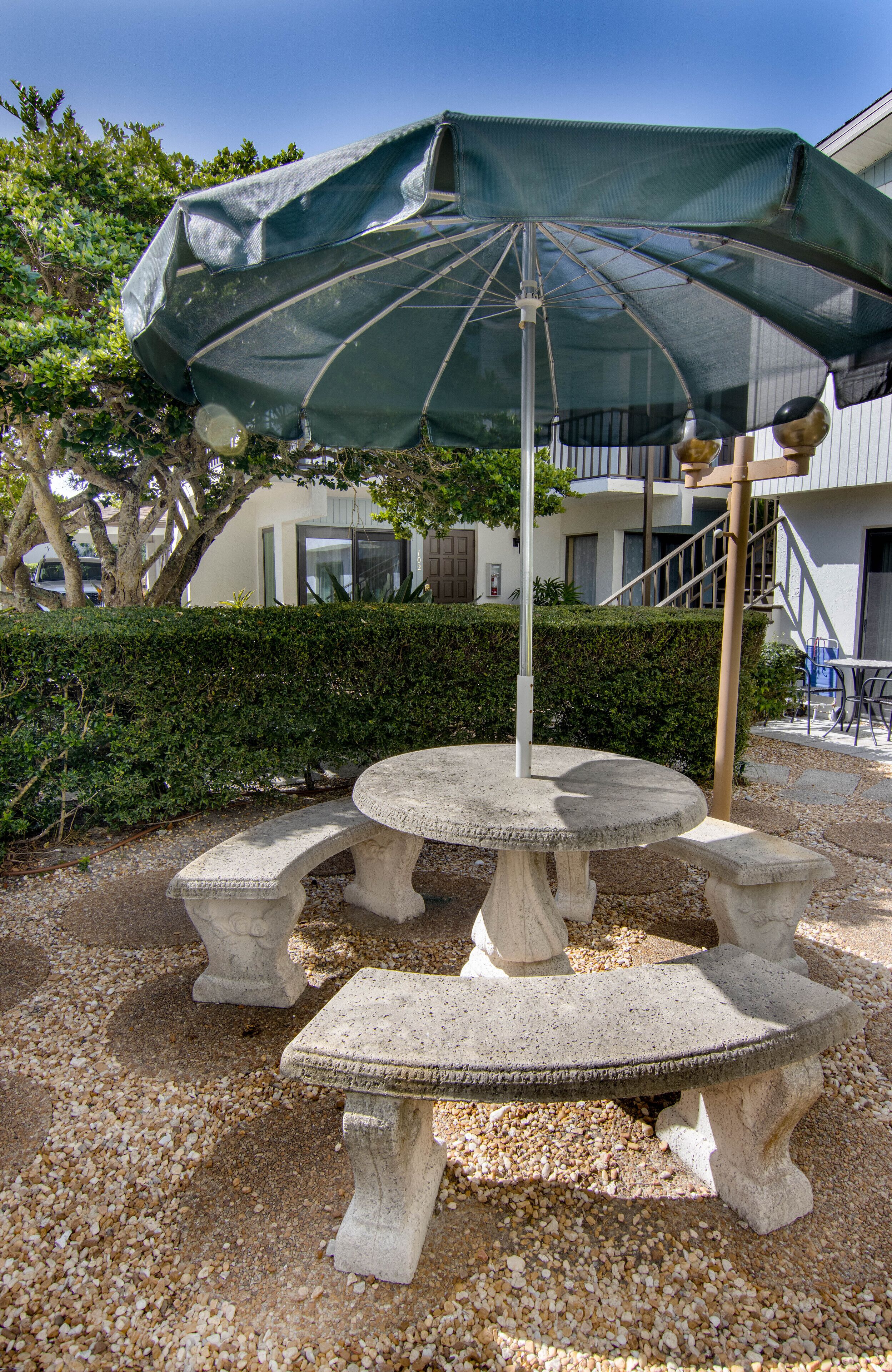 Un espace extérieur aménagé avec des sièges en pierre, une table et des bancs, un parasol, et un bâtiment en arrière-plan.