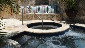 Un bain à remous avec un mur en pierre en arrière-plan, un coin salon avec des coussins à rayures et une plante en pot.