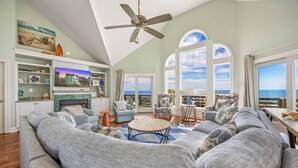 A spacious living room with a large gray sofa, a wooden coffee table, and a ceiling fan. There are multiple windows offering a view of the ocean.