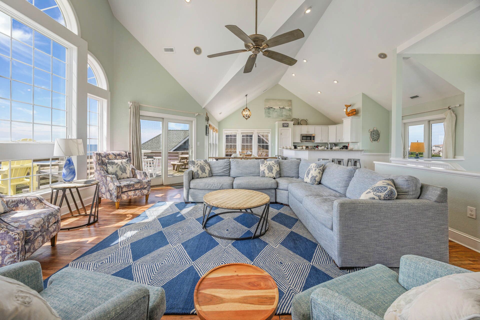 A spacious living room with a large sectional sofa, a round coffee table, and a patterned rug. There is a ceiling fan, a dining area with chairs, and a kitchen visible through an open door.