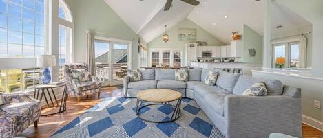 A spacious living room with a large sectional sofa, a round coffee table, and a patterned rug. There is a ceiling fan, a dining area with chairs, and a kitchen visible through an open door.