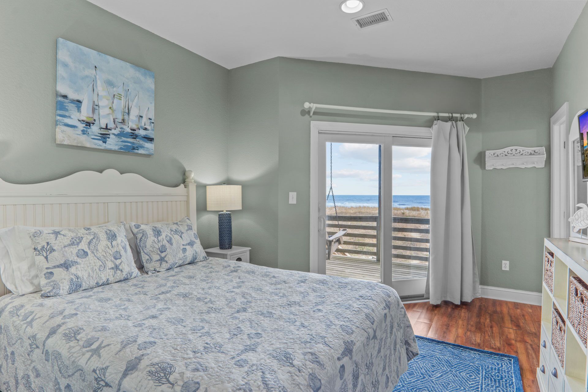 A bedroom with a bed, a painting of sailboats, a nightstand with a lamp, a view of the beach through a sliding door, and a dresser with storage drawers.