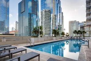 A rooftop pool with lounge chairs and palm trees, surrounded by high-rise buildings.