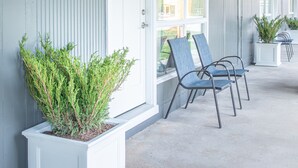 A covered porch with a row of chairs and potted plants, a white door, and a window with a view of the street.