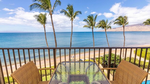 A balcony with wicker chairs and a glass table overlooking a beach with palm trees and a clear blue sea.