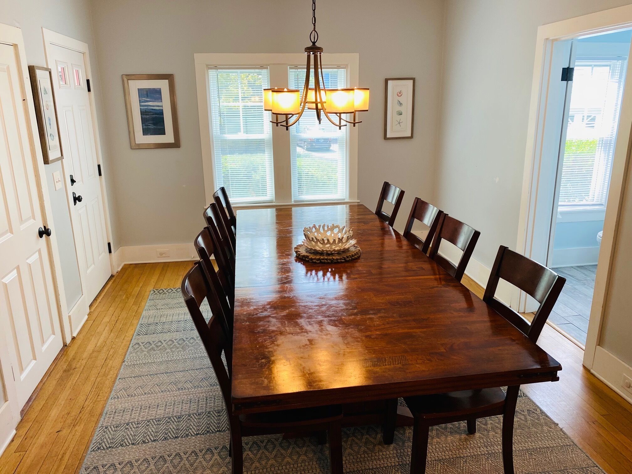 A dining room with a wooden table, eight chairs, a chandelier, and two windows with blinds.