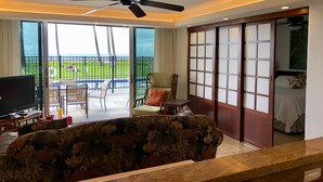 Habitación de hotel con un sofá grande, una televisión, un ventilador de techo, una puerta corredera y vistas a una piscina y zonas verdes.